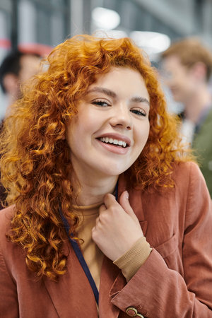 professional headshot of overjoyed redhead businesswoman looking at camera near blurred colleaguesの写真素材