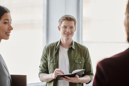 positive businessman writing in notebook near colleagues on blurred foreground in modern office