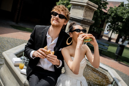 african american bride eating burger near redhead groom in sunglasses near fountain, outdoor weddingの写真素材