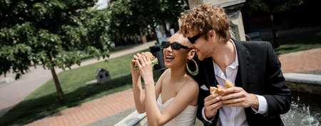 cheerful interracial newlyweds snack with burgers near city fountain, outdoor wedding, bannerの写真素材