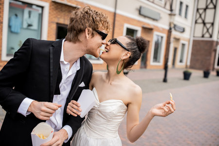 urban romance, outdoor wedding, multiethnic newlyweds in sunglasses eating french fries togetherの写真素材
