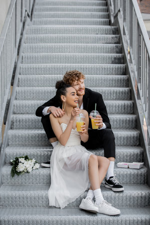 happy interracial newlyweds with orange juice looking away on street stairs near bouquetの写真素材