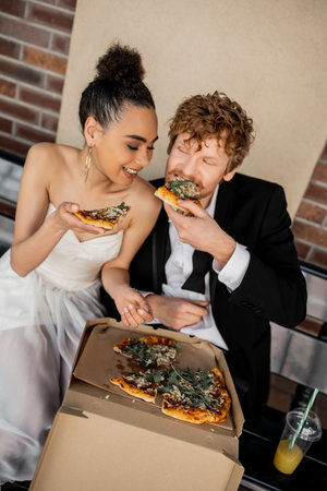 pleased multiethnic couple in wedding attire eating tasty pizza on bench, wedding in cityの写真素材