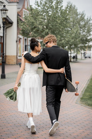back view of stylish interracial newlyweds with flowers and longboard embracing and walking in cityの写真素材
