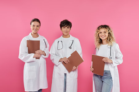 cheerful doctors holding paper folders while standing isolated on pink, breast cancer conceptの写真素材