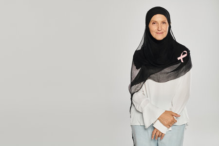 cheerful woman in hijab and pink ribbon of breast cancer awareness standing isolated on greyの写真素材