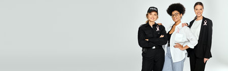 joyful interracial women with pink ribbons of breast cancer hugging isolated on grey, bannerの写真素材