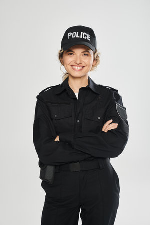 cheerful policewoman in uniform crossing arms and posing isolated on grey