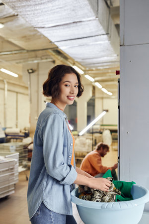 smiling young asian woman looking at camera near clothes in basket in blurred public laundryの写真素材