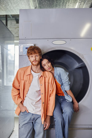 young asian woman sitting on washing machine near boyfriend in public laundryの写真素材