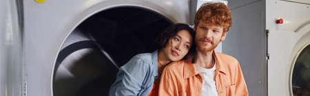 young asian woman standing near redhead boyfriend near washing machine in coin laundry, banner