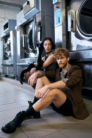 confident man in jacket sitting near asian girlfriend in dress and washing machine in public laundryの写真素材