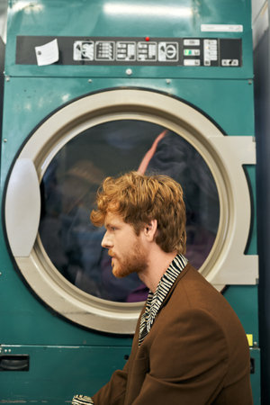 side view of fashionable young redhead man posing near washing machine in public laundryの写真素材