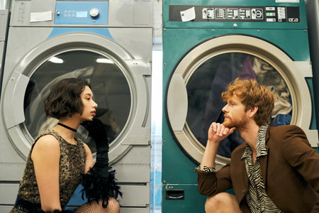 side view of trendy multiethnic couple looking at each other near washing machines in public laundryの写真素材