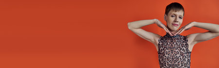androgynous person smiling and posing on orange backdrop, hands near face, queer fashion, banner
