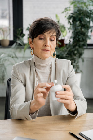 middle aged businesswoman holding earphone case with earbuds at workplace in modern officeの写真素材