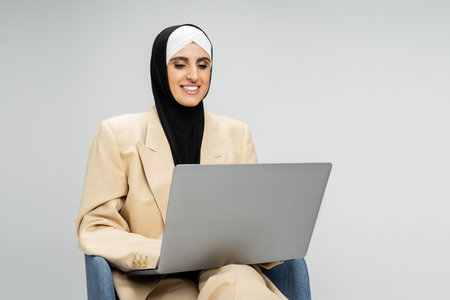 satisfied businesswoman in suit and hijab networking on laptop in armchair on greyの写真素材