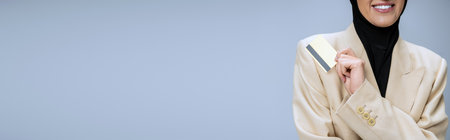 cropped view of smiling muslim business woman in blazer and hijab with credit card on blue, banner