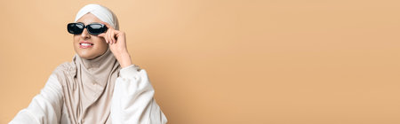 joyful muslim woman in hijab and trendy sunglasses looking away on beige, personal style, banner
