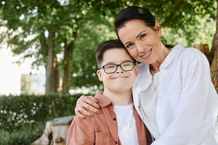 middle aged woman smiling at camera and hugging pleased son with down syndrome in parkの写真素材