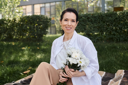 joyful middle aged woman in stylish clothes holding flowers and looking at camera on blanket in parkの写真素材