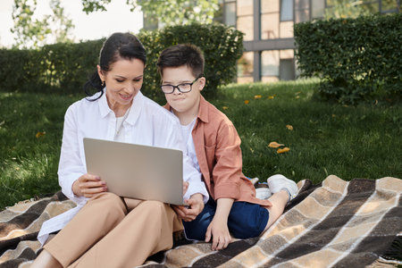 kid with down syndrome looking at laptop near smiling mother on blanket in park, e-learningの写真素材