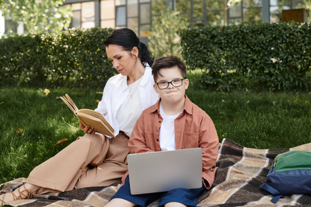 middle aged woman reading book near son with down syndrome and laptop on blanket in park, leisureの写真素材