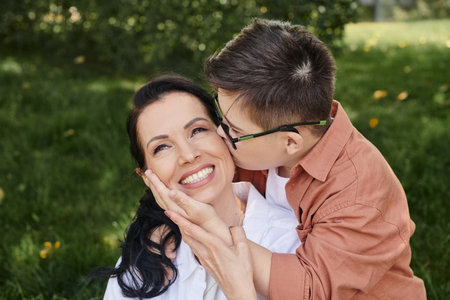 preteen boy with down syndrome, in eyeglasses, kissing overjoyed mother in park, unconditional loveの写真素材