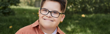 cheerful and genuine kid with down syndrome in eyeglasses smiling in park, portrait, banner