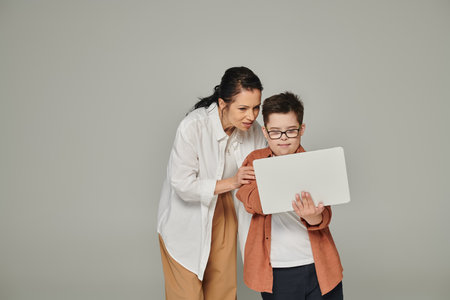 middle aged woman explaining online lesson to smiling son with down syndrome near laptop on grey