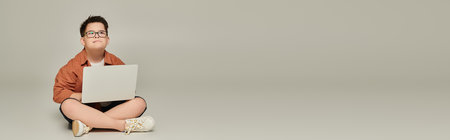 boy with down syndrome sitting with crossed legs and laptop while thinking on grey, banner