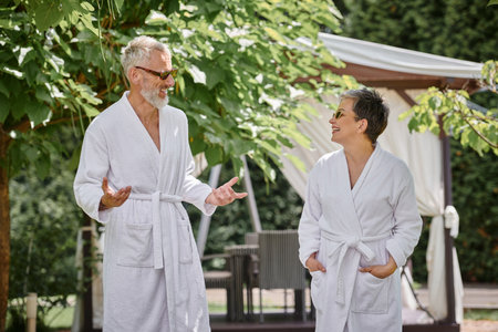 happy mature couple in sunglasses and robes chatting in luxury resort, wellness retreat conceptの写真素材
