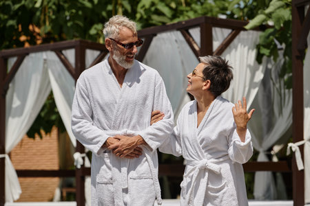 happy middle aged couple in sunglasses and robes walking in luxury resort, wellness retreat conceptの写真素材