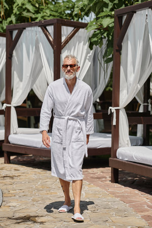 middle aged man in stylish sunglasses and white robe walking near private pavilion in luxury resortの写真素材
