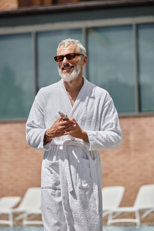 happy and tattooed middle aged man in sunglasses and robe holding smartphone in resort, retreatの写真素材
