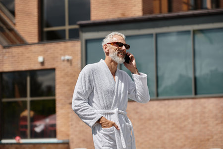 happy and tattooed middle aged man in sunglasses and robe talking on smartphone in resort, retreatの写真素材