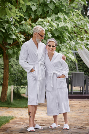happy mature man with tattoo hugging wife in sunglasses and robe in summer garden, wellness retreatの写真素材