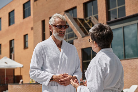 happy middle aged couple in sunglasses and robes chatting during vacation near hotel buildingの写真素材