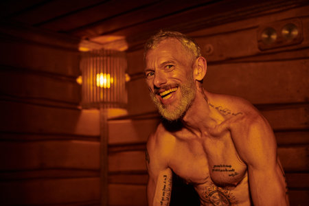 joyful and shirtless middle aged man with tattoos sitting in sauna, wellness retreat conceptの写真素材