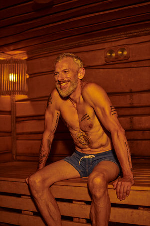 positive and shirtless middle aged man with tattoos sitting in sauna, wellness retreat conceptの写真素材