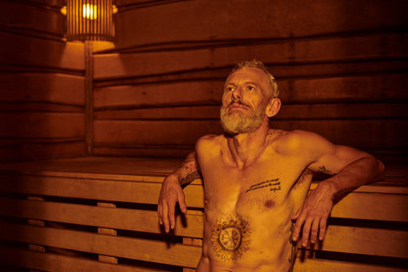 pensive and shirtless middle aged man with tattoos sitting in sauna, wellness retreat conceptの写真素材