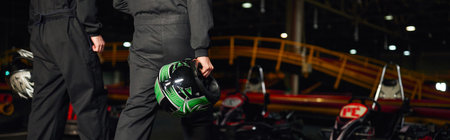 cropped view of go kart drivers in sportswear walking on circuit and holding helmets, banner