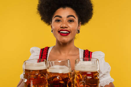 joyful african american bavarian waitress looking at camera near mugs with craft beer on yellow