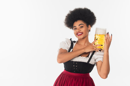 cheerful african american waitress in outfit holding mug of beer with foam on whiteの写真素材