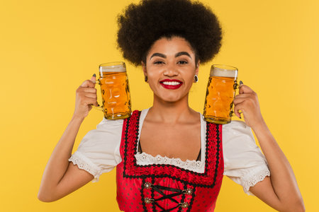 smiling african american waitress in dirndl dress holding mugs with craft beer on yellowの写真素材