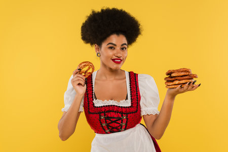 joyful african american waitress in authentic costume holding pretzels on yellowの写真素材