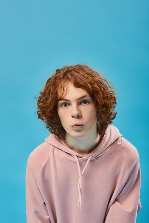 portrait of impressed teenage boy with red wavy hair in stylish hoodie and looking at camera on blue