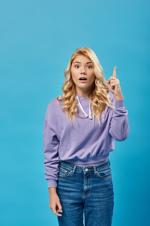 inspired blonde teenage girl with open mouth showing idea gesture and looking at camera on blueの写真素材