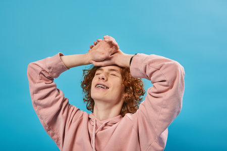 positive, dreamy redhead teen guy in hoodie standing with closed eyes and hands on forehead on blue