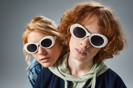 portrait of redhead teenage guy and girlfriend in fashionable sunglasses looking at camera on greyの写真素材